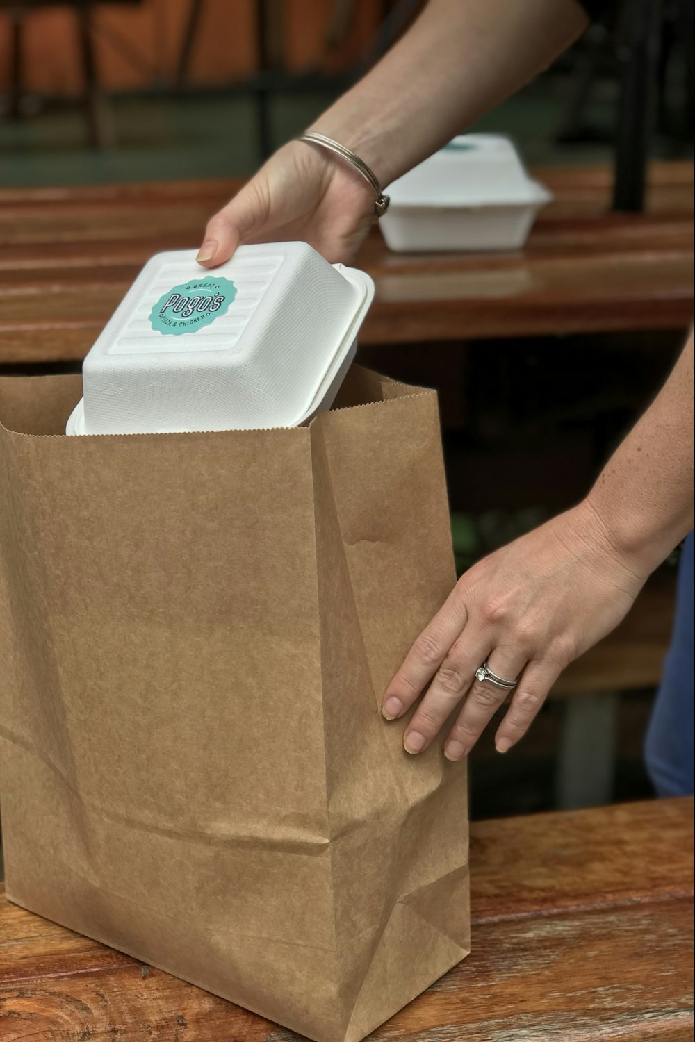 Hands packing a takeaway container into a brown paper bag.